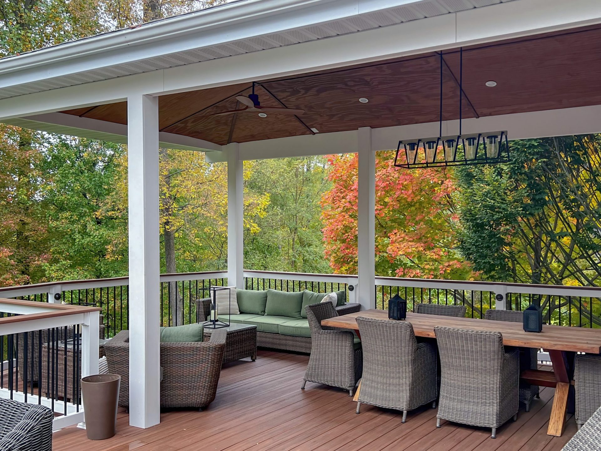 Covered deck with a wood paneled ceiling, custom outdoor lighting, and quality comfortable dining and lounge furniture overlooking autumn trees in Northern New Jersey.