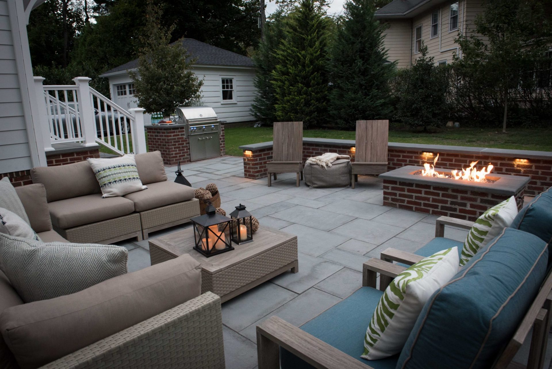 A bluestone patio with a rectangular brick gas fire pit, integrated under-cap retaining wall lighting, and a built-in stainless steel grill station in a backyard.
